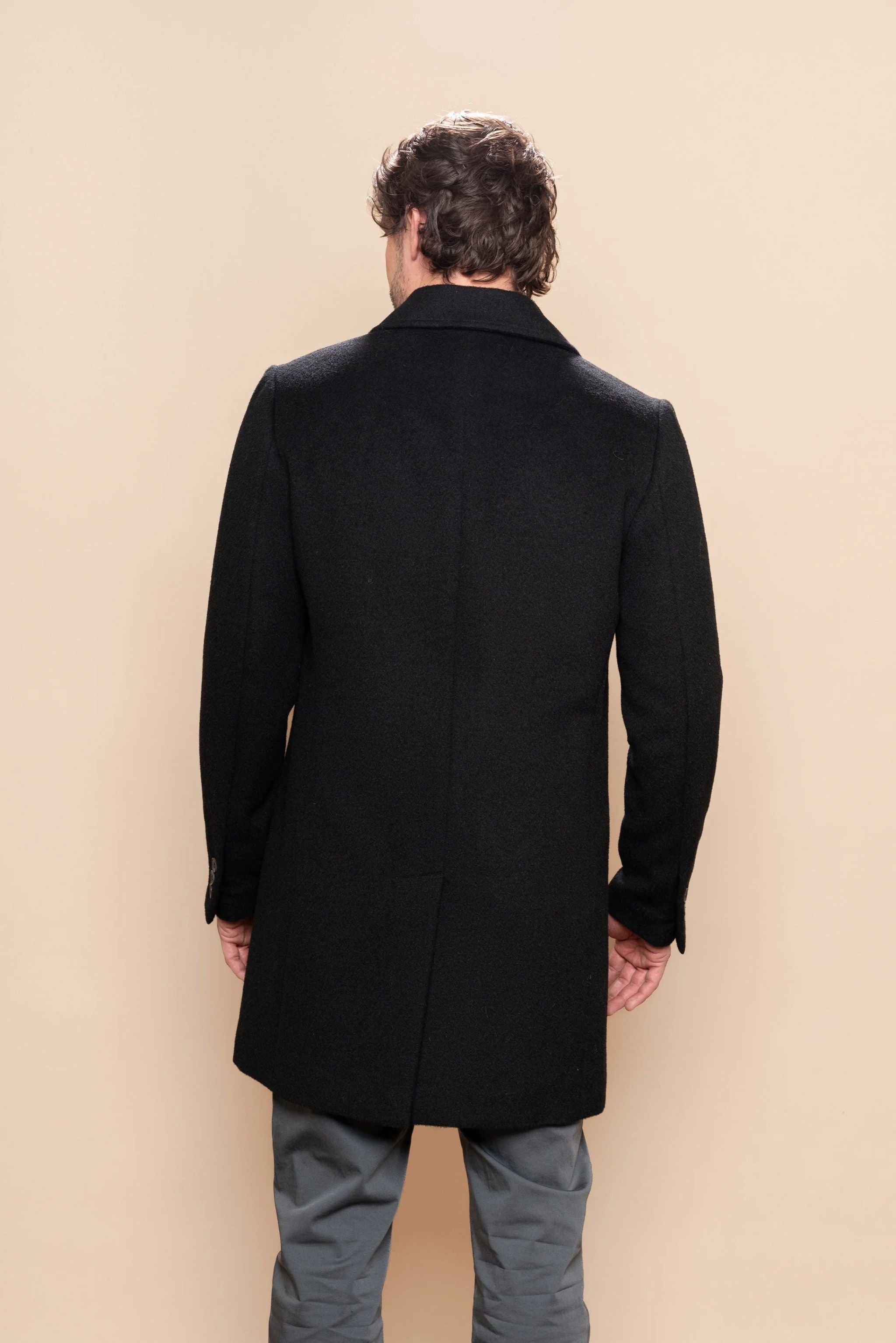 Man wearing a black coat standing against a beige wall